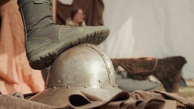 Low-angle close-up shows boot pressing down on antique metal helmet inside canvas tent, with clear contact, slight indentation, riveted patina, warm light, soft shadows, and shallow focus