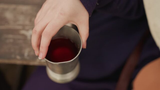Overhead closeup of adult hand tilting metal cup filled with dark red liquid, showing careful pouring action near rim, warm metallic reflections, and soft light over intimate tabletop scene