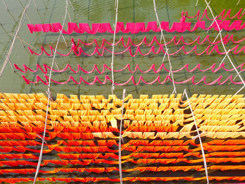 Aerial view of vibrant red, orange, and yellow fabrics drying on bamboo frames over water with workers tending to the textiles in Narayanganj, Dhaka, Bangladesh.