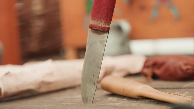 Low angle view shows knife blade pressed into wooden tabletop beside wooden spoon and folded cloth, with warm ambient light, shallow focus, soft blur, and blurred pottery in background
