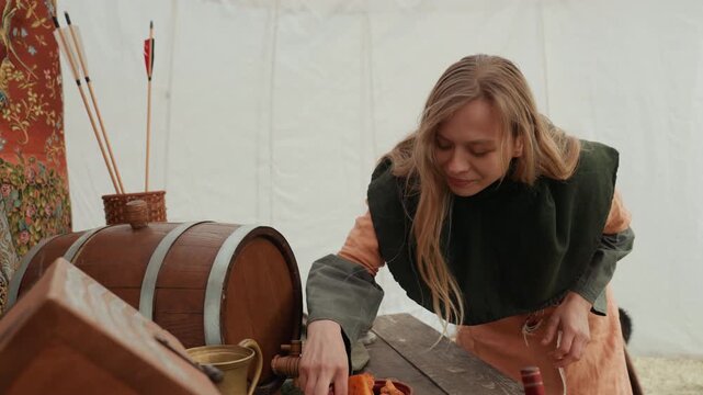 Adult woman studies small objects on wooden table inside canvas tent, leaning forward with focused expression, arranging items carefully before standing and moving away in camp setting