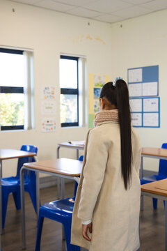 African American teen female standing wearing beige coat and scarf in classroom with blue chairs