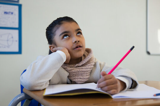 Teenage girl sitting at school desk, resting elbow, holding bright pink pencil above open notebook