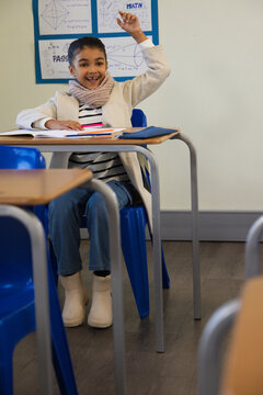 School-age female student sitting at school desk raising right hand smiling with notebook and pens