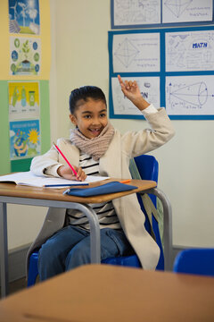 Female child sitting at school desk writing with red pencil over open notebook, wearing beige coat