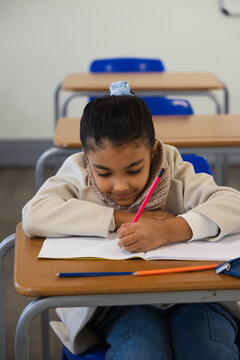 Youth student at school desk wearing beige coat and scarf writing in notebook holding pink pencil