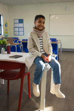 African American child sitting on table edge in classroom wearing tan coat with notebook and bottle