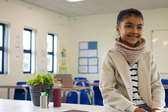 African female child sitting front-right classroom wearing beige coat near plant laptop, copy space