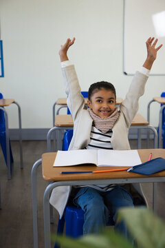 African American child sitting at front desk on blue chair, raising arms, smiling, open notebook