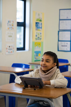 African American schoolgirl wearing coat and knitted scarf, sitting at desk using tablet on stand