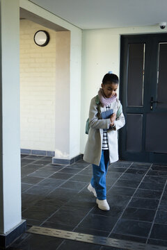 African American teenage female standing in foyer in coat holding tablet and backpack, copy space