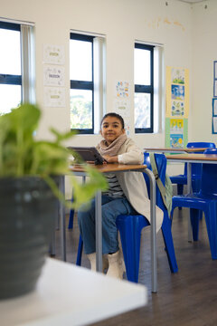 African American schoolchild sitting at desk in classroom using tablet with backpack hanging