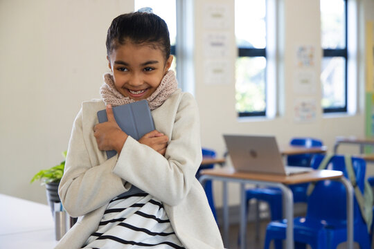 Teen girl sitting on desk edge wearing coat, scarf hugging tablet with case in classroom smiling
