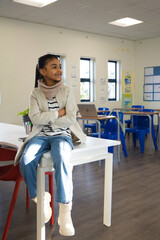 Fototapeta premium African female child sitting on white rectangular table in classroom at school wearing beige coat