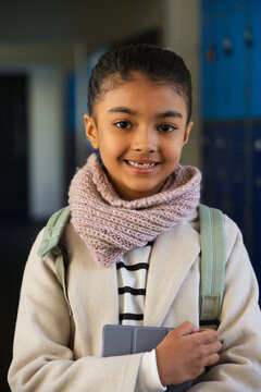 African American child standing in school hallway holding gray tablet wearing backpack and scarf