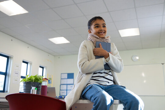 African teenage girl sitting on desk edge holding grey tablet in classroom, wearing coat and scarf
