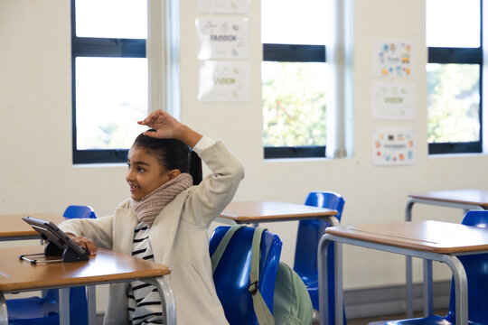 Youth woman of African descent in coat sitting at classroom desk raising hand using tablet backpack