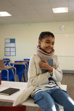 Female child sitting on white table at front of classroom wearing beige coat clutching gray tablet