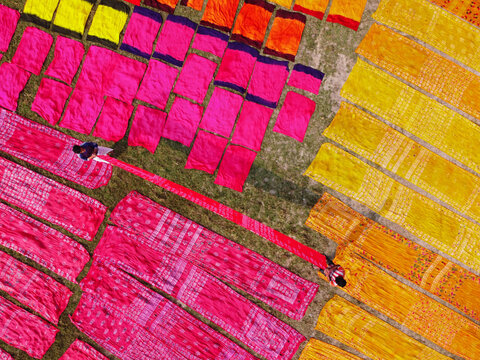 Aerial view of vibrant pink, orange, and yellow fabrics laid out to dry on a grassy field with workers tending to the textiles in Narayanganj, Dhaka Division, Bangladesh.