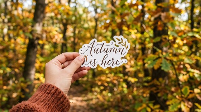 Woman holding sticker with text autumn is here outdoors in scenic forest surrounded by yellow leaves