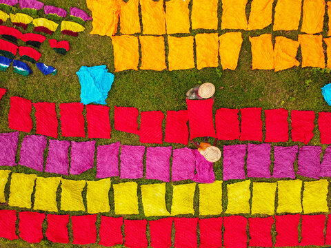 Aerial view of colorful fabrics drying on the grass with workers wearing traditional hats in Narayanganj, Dhaka Division, Bangladesh.