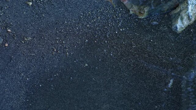 Clear shallow lake water ripples over dark gravel near shoreline rocks, revealing fine sediment and scattered pebbles beneath transparent freshwater along the natural edge of a mountain lake