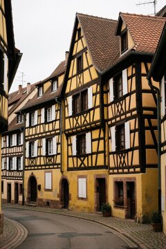 Picturesque street in Dambach la Ville, Alsace, features historic half timbered buildings with vibrant yellow facades, dark wood beams. Cobblestone pavement lines quiet village lane. Old European