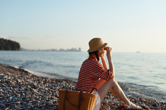 A woman on a pebbled beach wears a striped shirt, straw hat, and carries a woven basket, enjoying a calm seaside moment under warm light.