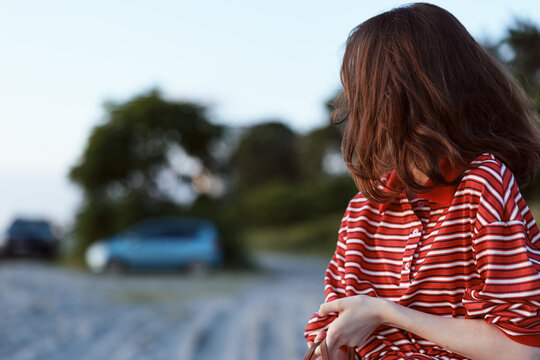 A young woman with brown hair standing along the shoreline wears a red striped shirt and looks down with a calm, thoughtful expression, while distant cars blur in the background.