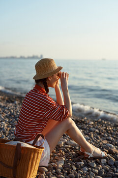 Woman sits on a pebble beach in a striped shirt and straw hat, gazing at the sea with a thoughtful pose. Soft golden light captures a calm summer moment by the water.