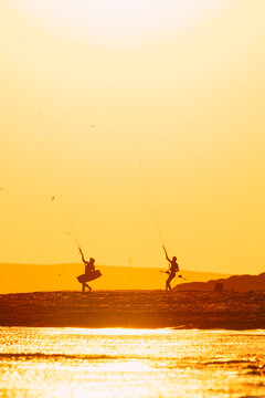 Two kite surfers on beach as dark silhouettes at golden sunset