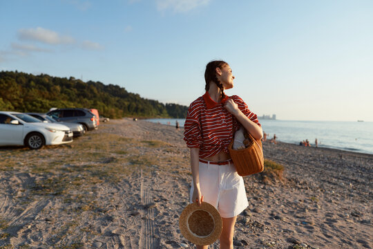One young woman in a stripedtop and white shorts walks along a sunny beach at golden hour, holding a hat and bag with a gentle breeze.