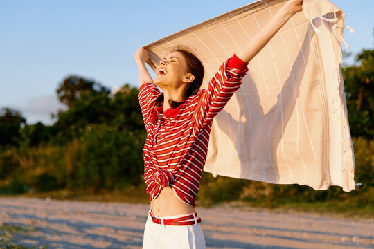 A young woman on a sunny beach laughs with arms raised, holding a striped towel like a sail, capturing carefree freedom and playful summer vibes.