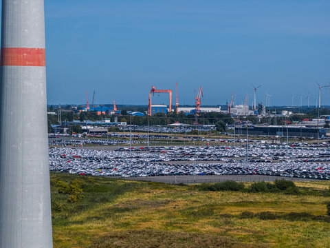 Aerial view of the Volkswagenwerk Emden car factory with a vast parking lot filled with new vehicles, industrial cranes, and wind turbines in Emden, Niedersachsen, Germany.