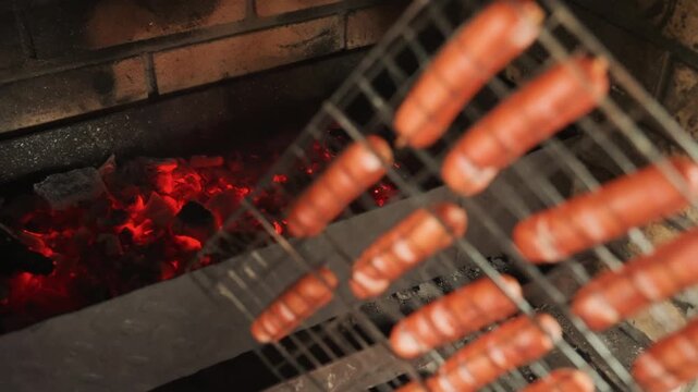 Close view of multiple sausages arranged in parallel rows cooking over glowing charcoal embers inside brick lined hearth, with active browning, sear marks, light smoke, and rustic warmth