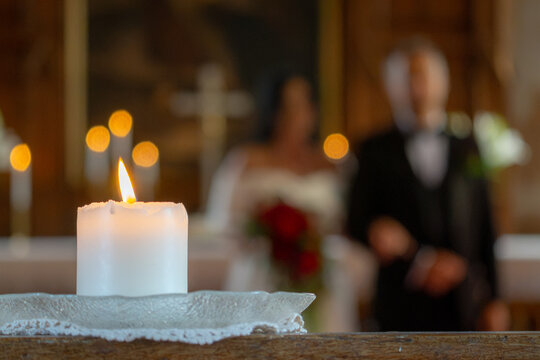 Burning white candle in focus with a blurred bride, groom and officiant during a church wedding ceremony