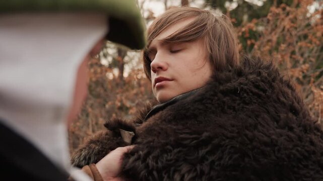 Teenage boy stands in woodland while peer secures dark fur-like cloak over shoulders, hands moving carefully as boy tilts head with subtle smile, calm acceptance, warm daylight, quiet ritual mood