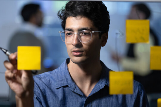 Indian male professional attaches a sticky note to a glass board while coworkers collaborate in the background during a brainstorming meeting in an office