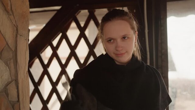 Young woman with braided hair leans forward to receive mug from person outside view, holding soft eye contact and calm expression beside textured lattice, wood, and stone inside historic room
