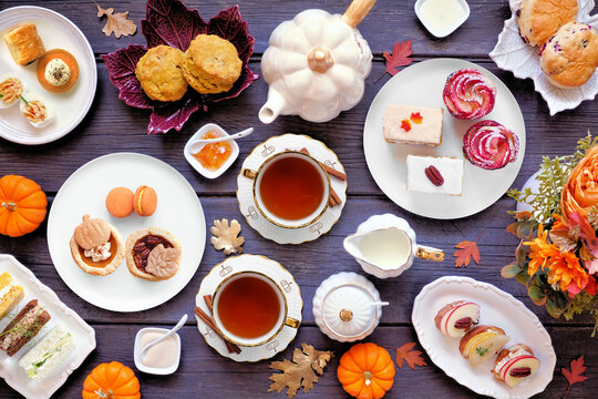 Fall theme afternoon tea table scene against a dark wood background. Pumpkin and apple theme savory finger foods, sandwiches, scones, and desserts. Top down view with copy space.