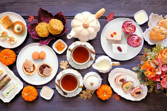 Autumn theme afternoon tea table scene against a dark wood background. Pumpkin and apple theme savory finger foods, sandwiches, scones, and desserts. Above view with copy space.