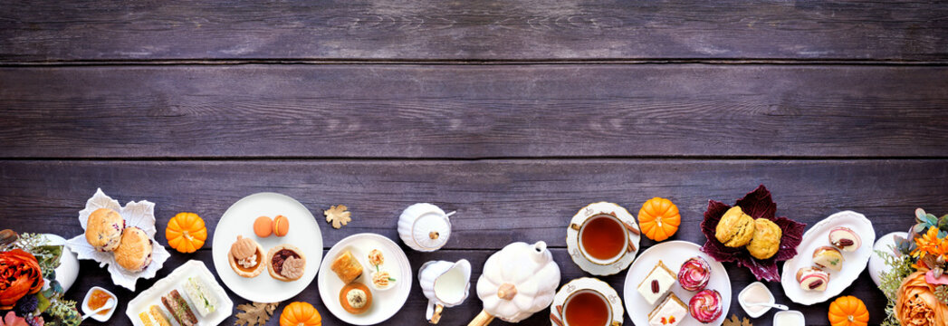 Fall theme afternoon tea bottom border against a dark wood banner background. Pumpkin and apple theme savory finger foods, sandwiches, scones, and desserts. Top view with copy space.