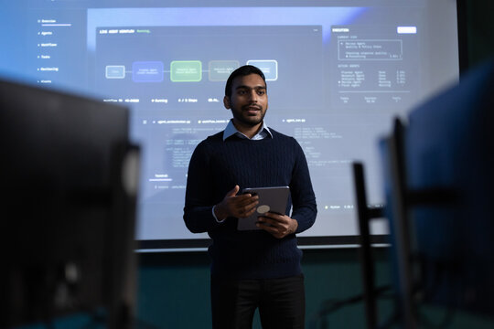 Young Indian male professional holds a tablet while presenting a software workflow to coworkers in a meeting room, with computer screens and listeners in the foreground.