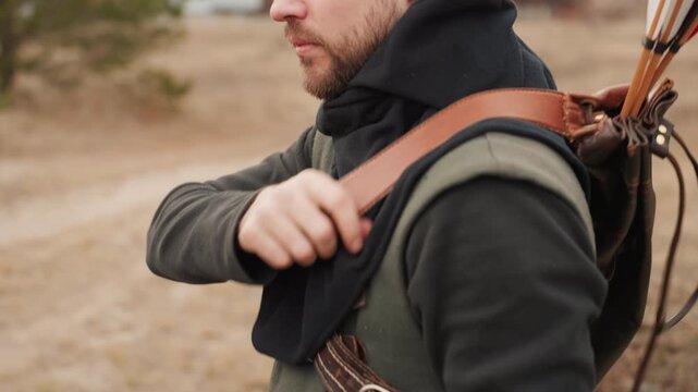 Profile close-up of male archer adjusting leather quiver strap over shoulder, hand gripping strap while torso turns slightly and gaze stays off-frame, showing readiness in soft daylight