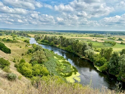 High-angle panoramic view of a calm winding river flowing through a lush green valley with golden grass on the hill under a cloudy blue sky.