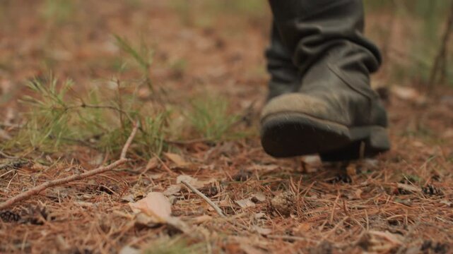 Close low angle view shows booted feet walking across dry pine needle covered forest ground, one boot planted with tread contact while other lifts mid step, emphasizing natural forward movement