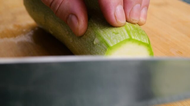 A chef uses a sharp knife to slice a fresh green zucchini into smooth round pieces on a wooden cutting board, preparing healthy organic ingredients for a home meal.