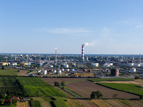 Aerial view of the PKN Orlen refinery complex with storage tanks, industrial pipes, and a tall chimney emitting smoke, surrounded by agricultural fields in Plock, Masovian Voivodeship, Poland.
