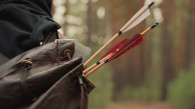 Rear view of hooded archer moving through dense pine woodland, wearing textured fabric cloak and strapped leather quiver with wooden arrows, showing calm focused outdoor motion in soft daylight