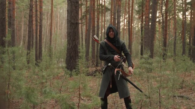 Hooded archer steps and pivots through dense pine woodland while raising bow arm and preparing to draw or release arrow, with visible torso rotation, forward foot placement, and tense focused posture